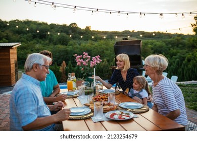 Group Of Family Members Enjoying A Dinner Party Out In The Yard. They Are Siting At The Dining Table And Eating