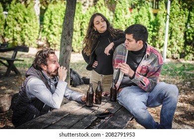 Group Of Female And Male Young Friends Drinks Beer From Bottle And Smoking Cannabis Marijuana Ganja Or Hashish Joint In Public Park Outdoor. Young People Smoke The Grass. Real Life Scene Concept