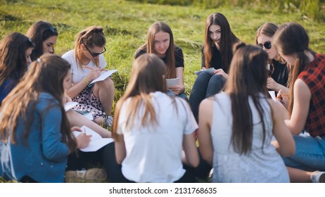 A Group Of Female Students Are Sitting In A Circle On A Meadow For Collective Work With Notebooks.