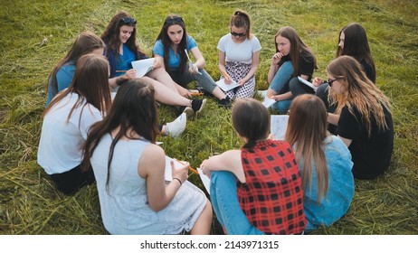 A Group Of Female Students Are Sitting In A Circle On A Meadow For Collective Work With Notebooks.