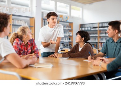 Group of fifteen-year-old schoolchildren are preparing for classes in the school library, discussing something and making notes in copybooks