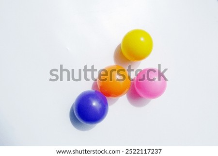A group of five brightly colored plastic balls arranged on a white surface. The balls are yellow, orange, pink, and blue. They are casting shadows on the white background.
