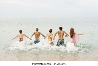 Group Of Five Friends Holding Hands And Running Into The Sea Water Together, Being Spontaneous And Having Fun While On A Summer Vacation On A Beach With Blue And Turquoise Waters.