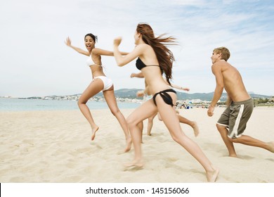 Group Of Five Friends Running Together Towards The Sea Water Shore, Being Spontaneous And Having Fun While On A Summer Vacation On A Beach Showing Happy Expressions.