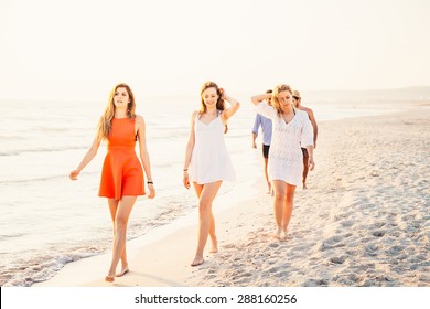 A Group Of Five Friends Walking On The Beach At Sunset. Two Young Men And Three Young Women In Small Groups, They Leave Behind The Sun And Walking On The Sand In A Day Of Rest.