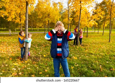 Group of five kids play hide and seek with boy counting and friends hiding behind the trees