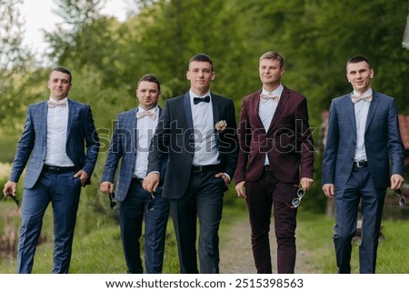 A group of five men are dressed in suits and ties, walking down a path. They are all wearing ties and one of them is holding a cigarette. Scene is formal and elegant, as the men are dressed in suits