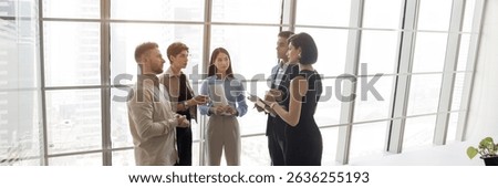 Group of five processionals discussing work-related matters gathered in modern office standing against panoramic window with skyscraper city view, wide copy-space image. Business interaction, teamwork