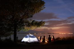Group Of Five Travellers, Girls And Boy Having A Rest On Lake Shore Under Tree Around Campfire At Tourist Tent On Quiet Water Surface And Blue Evening Sky Background. Tourism And Camping Concept.