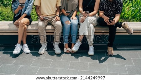 Group Of Five Unrecognizable Multi Ethnic Friends Using Mobile Phones and Having Fun Outdoors. 
Cropped photo of friends watching or typing something on their phones while sitting in a row on a bench.