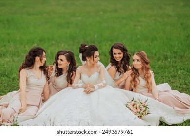 A group of five women are sitting on the grass, with one of them wearing a wedding dress. The bride is surrounded by her bridesmaids, and they are all smiling and enjoying each other's company - Powered by Shutterstock