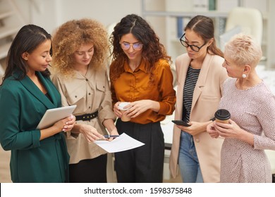 Group of five women talking about work during coffee break standing together in office room