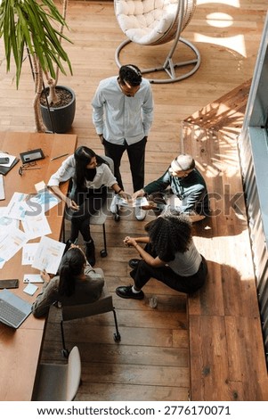 A group of five workers stand and sit on a bench and chairs at a table while looking at a document that two of them are showing