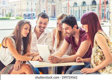 A group of five young friends (two men and three women) looking at a tablet sitting together at a table in a bar in the arcades in a hot summer day - Powered by Shutterstock