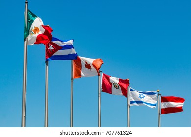 A group of flags waving on flag poles from the countries of Austria, Canada, Cuba, Israel, Mexico, and Peru.