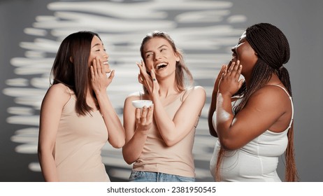 Group of flawless ladies applying moisturizing face cream in studio, promoting luminous skincare products. Interracial young women using moisturizer and serum for new dermatology campaign. - Powered by Shutterstock