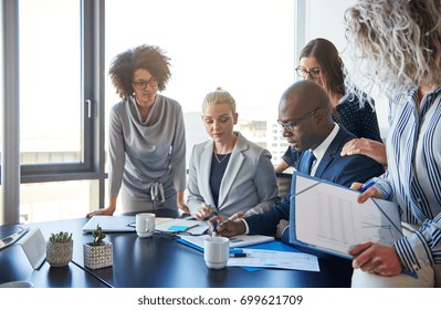 Group of focused businesspeople standing around their manager sitting at an office boardroom table reviewing charts and paperwork