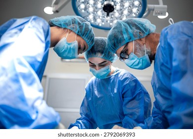 Group of focused surgeons looking down at patient during operation, healthcare and medicine concept. Team of professional medical specialists performing deep surgical procedure under hospital light.