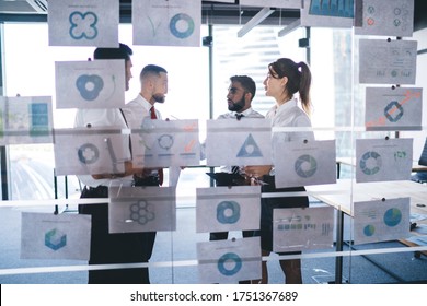 Group Of Formally Dressed Workers Discussing Business Diagrams Placed On Glass Wall While Standing In Modern Open Space Office With Panoramic Windows