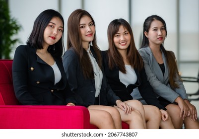 Group Of Four Attractive Asian Female Office Colleagues In Formal Business Suits Sitting On Red Couch In Office Smiling At Camera. Concept For Modern Office Working.