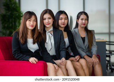 Group Of Four Attractive Asian Female Office Colleagues In Formal Business Suits Sitting On Red Couch In Office Smiling At Camera. Concept For Modern Office Working.