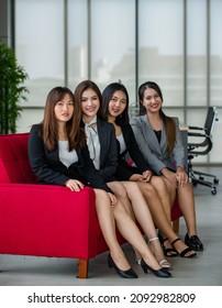 Group Of Four Attractive Asian Female Office Colleagues In Formal Business Suits Sitting On Red Couch In Office Smiling At Camera. Concept For Modern Office Working.