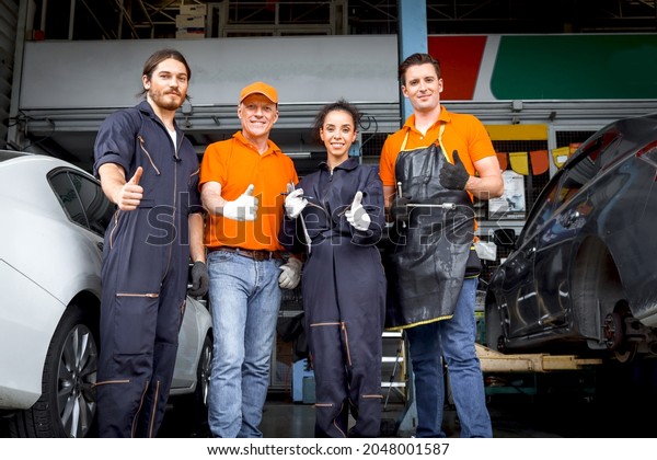 Group of four car service technician men and woman\
giving thumbs up to camera, people working together at vehicle\
repair garage service shop, check and repair customer car at\
automobile service center