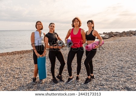 Group of four Caucasian pretty adult women holds sports mats on pebble beach. Sea and wild coast in background. Fitness outdoor training.