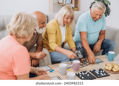 Group of four cheerful senior people, two men and two women, having fun sitting at table and playing bingo game in nursing home