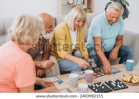 Group of four cheerful senior people, two men and two women, having fun sitting at table and playing bingo game in nursing home