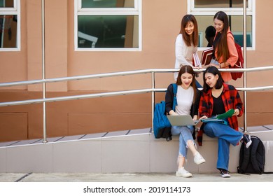 Group Of Four College Student Girls Holding Books And Notebook Computer Sitting And Talking Together With Intimate In Front Of School Building. Learning And Friendship Of Teens Close Friend Concept.