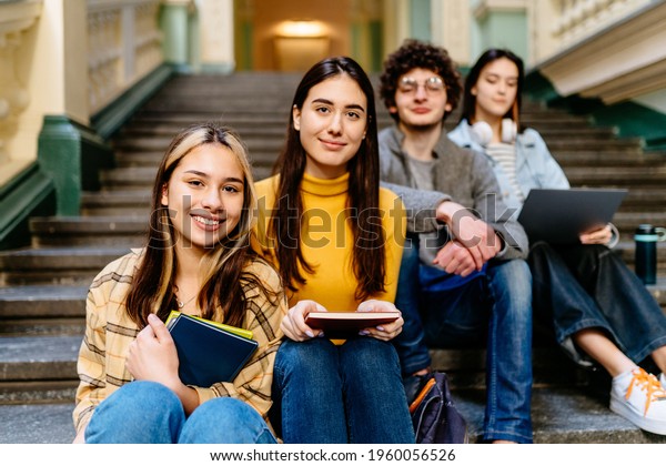 Group of four college students at the\
university or college looking happy. Multinational team. A three\
women and a young curly man sitting on the stairs of the university\
for teamwork.