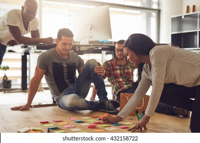 Group Of Four Employees Meeting Around Various Colored Sticky Notes On Hardwood Floor In Small Office