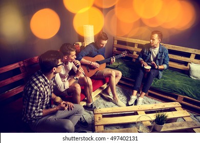 Group Of Four Friends Hanging Out In Studio. Guys Singing And Playing Music In Urban Apartment. Toned Photo With Bokeh.