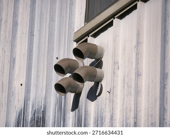 A group of four gray exhaust vent pipes protruding from a corrugated metal industrial wall under bright sunlight.