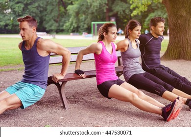 Group of four male and female adults performing dip exercises on park bench outdoors during summer - Powered by Shutterstock