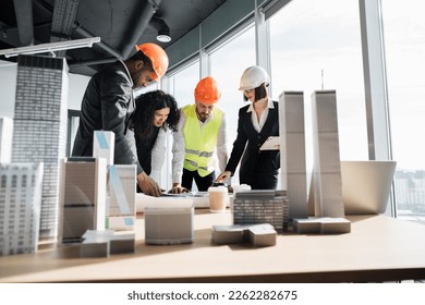 Group of four multiracial people architects standing near desk with lots of blueprints and architecture design of buildings with residential project maquette at office with panoramic windows. - Powered by Shutterstock
