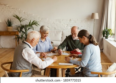 Group Of Four Senior Friends, Two Men And Two Women, Sitting At Table And Playing Bingo Game In Assisted Living Home