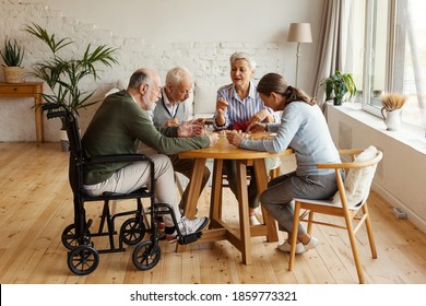 Group Of Four Senior People, Two Men Including Disabled One And Two Women, Sitting At Table Together And Playing Bingo Game In Nursing Home