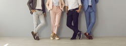 Group Of Four Successful Company Leaders In Classy Formal Suits Standing Hand In Pocket. Team Of 4 Business Partners Leaning On Grey Office Wall. Cropped Shot Of People's Legs In Stylish Classic Pants