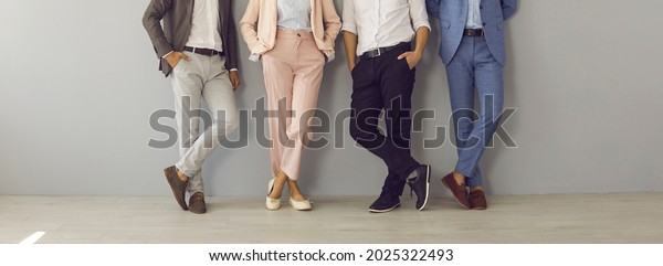 Group of four successful company leaders in classy\
formal suits standing hand in pocket. Team of 4 business partners\
leaning on grey office wall. Cropped shot of people\'s legs in\
stylish classic pants