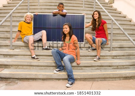 Group of four teenagers of mixed ethnicities sitting on a staircase and posing with a solar panel to promote the use of alternate sources of energy