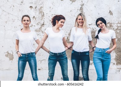 Group of four young diverse girls wearing blank white tshirt and jeans posing against rough street wall, fashion urban clothing style, mockup for t-shirt print store
