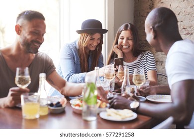 Group Of Four Young Friends Having Dinner In Cozy Cafe While Women Sharing New On Phone. 