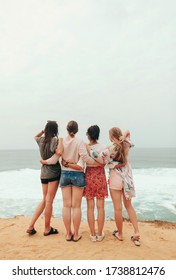 Group of four young stylish cuddiling girls looking at the ocean waves view on the beach in summer vacation. Women Friendship. Back view. Copy space