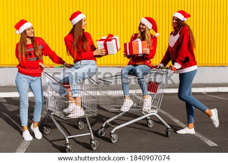 Group of four young women in christmas sweaters and santa claus hats with christmas gifts on carts near shopping mall