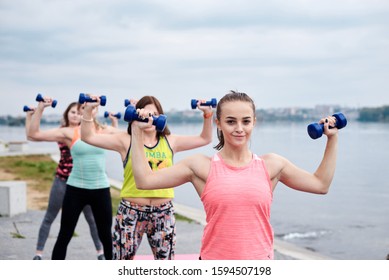 A Group Of Four Young Women, Wearing Colorful Sports Outfit,doing Fitness Exercises With Small Weights Outside By Lake In Summer.Workout Power Female Training To Loose Weight And Body Shape At Nature.