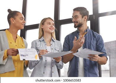 Group of friendly three multiethnic business people stand in office hall, hold papers, study financial report, try come to solution how clear debts, have pleased expressions as going to have corporate