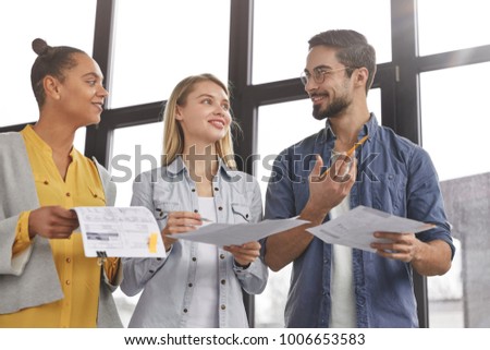 Group of friendly three multiethnic business people stand in office hall, hold papers, study financial report, try come to solution how clear debts, have pleased expressions as going to have corporate