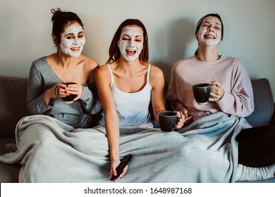 
Group Of Friends Applying A Revitalizing White Mask On Their Faces. Beauty Treatment And Skin Care. Seated On The Sofa Drinking Coffee And Watching Tv Together. Lifestyle.